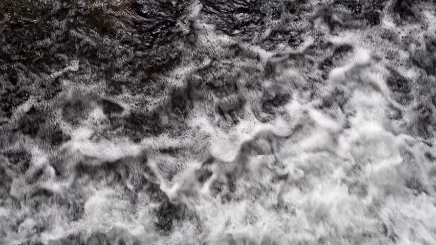 Overhead close up view of turbulent river water surface flowing rapidly over a dam. Rushing stream creating white foam, bubbles, and abstract textures. Concept of hydro energy, power, and nature background.