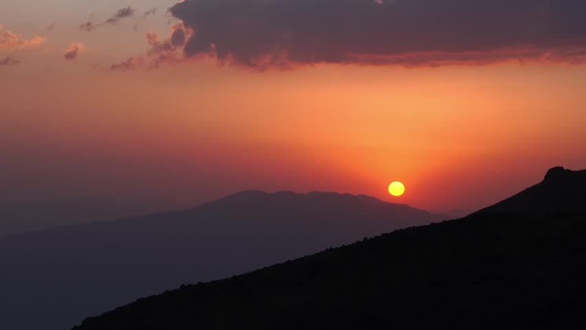 Fiery sun setting behind mountain ridges seen from Mount Ararat base camp, dramatic sky with surreal glow and symbolic solitude mood