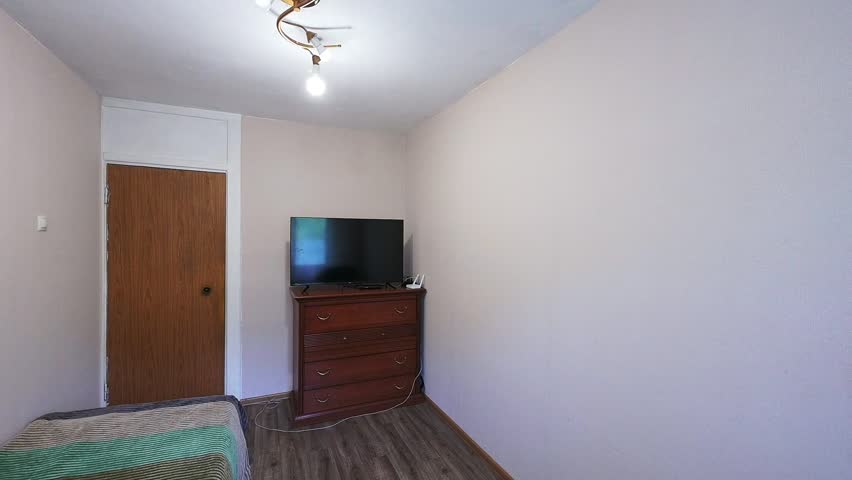 simple room with a wooden door, chest of drawers with a TV on top, a bed partially visible, plain walls, and a basic ceiling light fixture. Minimalist setup, neutral tones