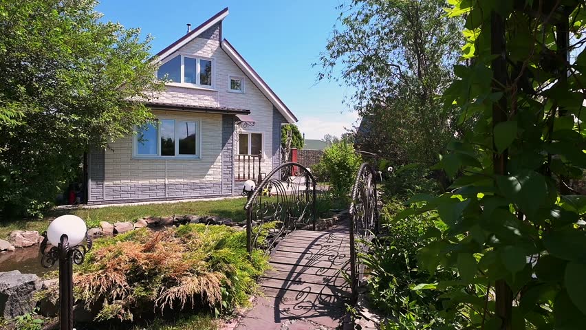 two-story house with light siding and large windows, surrounded by greenery. A decorative metal bridge crosses a small pond, leading to the house. Trees and lawn enhance the serene setting