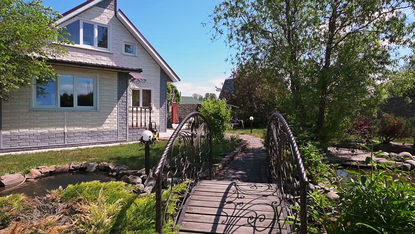 charming house with a wrought iron bridge over a small pond, surrounded by lush greenery, trees, and a well-maintained garden path leading to the entrance