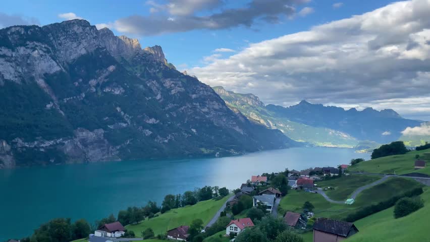 Panoramic view of Walensee lake and Churfirsten Mountains in Switzerland. Turquoise water and green alpine slopes with traditional houses under a blue sky with clouds. 