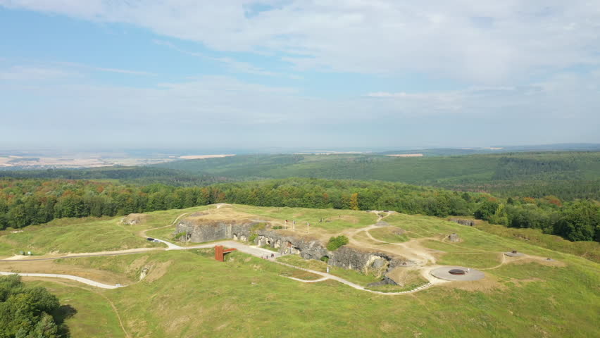 Wide aerial shot of Fort de Douaumont surrounded by green fields and forest under a partly cloudy sky in France.