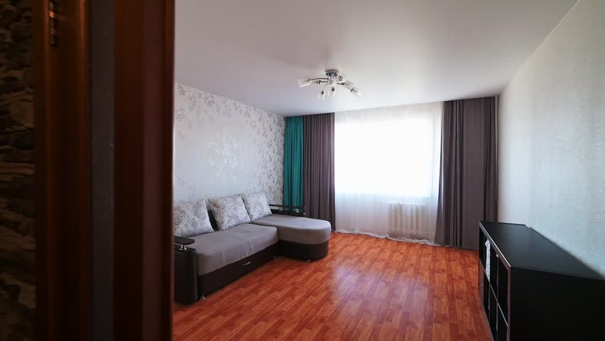 A cozy living room with a grey sectional sofa, floral-patterned cushions, light wallpaper, wooden flooring, and layered curtains — bright and welcoming