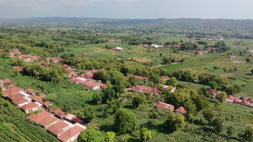 A panoramic aerial view of a rural village with houses featuring red-tiled roofs nestled among lush green trees and expansive agricultural fields.