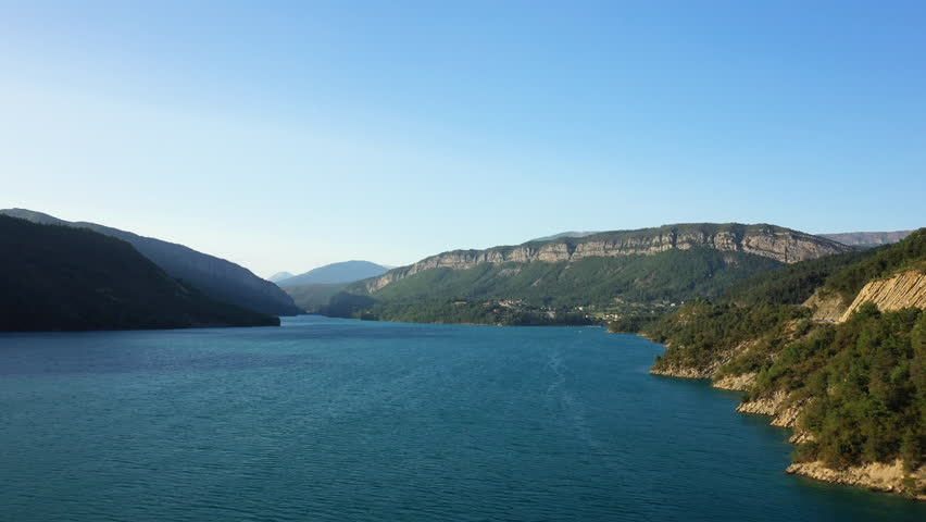 Wide view of Lac de Castillon with turquoise water surrounded by lush green mountains under a clear blue sky in southeastern France.