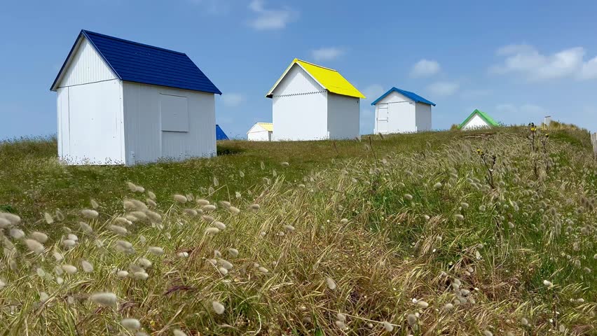 Colourful Beach Cabins in dunes at Gouville-sur-Mer, Normandy, France. Atlantic ocean. Colorful beach cabins with red, blue, and yellow roofs stand amidst sandy dunes and sparse grass. Their walls are painted white, contrasting sharply with the vibrant roofs. The clear blue sky with scattered clouds creates a picturesque backdrop in Normandy, France.