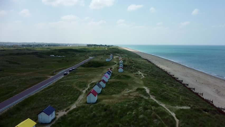 Aerial photo of Colourful Beach Cabins in dunes at Gouville-sur-Mer, Normandy, France. Atlantic ocean. Colorful beach cabins with red, blue, and yellow roofs are scattered across grassy dunes in Gouville-sur-Mer, Normandy. A sandy path meanders through the dunes, leading down to the Atlantic Ocean. The sea appears calm under a lightly clouded blue sky. In the distance, a car is parked near the cabins, and a few houses are visible, blending with the natural scenery. The idyllic coastal landscape showcases a mix of nature and human-made structures.