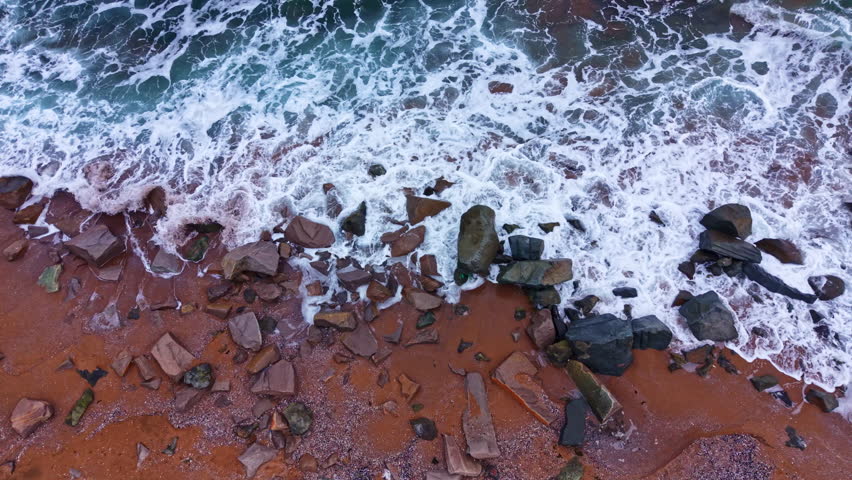 Waves move onto a rocky shore with sand during daytime. The water splashes against the stones, creating a natural scene by the ocean.