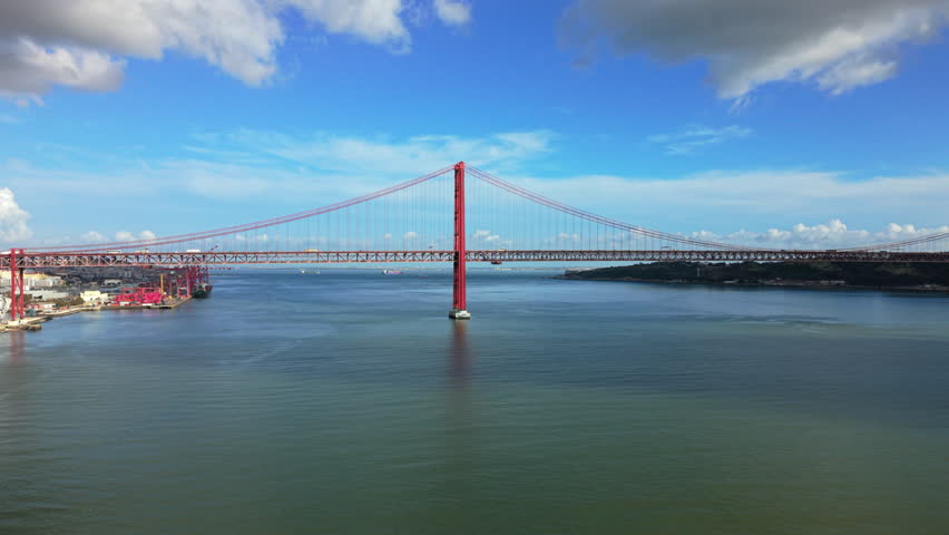 Aerial panorama of Lisbon Portugal showing 25 de Abril Bridge over Tagus River, wide water surface, distant riverbanks and urban shoreline under blue sky.