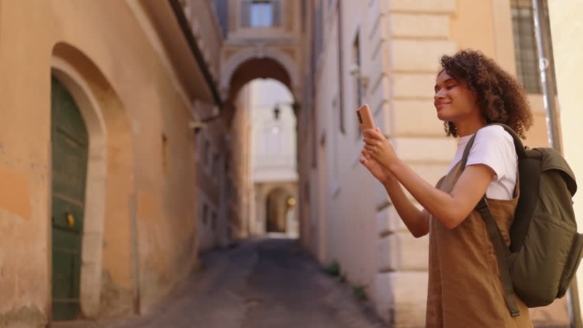 Smiling female traveler with a backpack holds her smartphone and taking a pictures while enjoying a walk through a narrow city street, capturing moments during a relaxed urban trip.