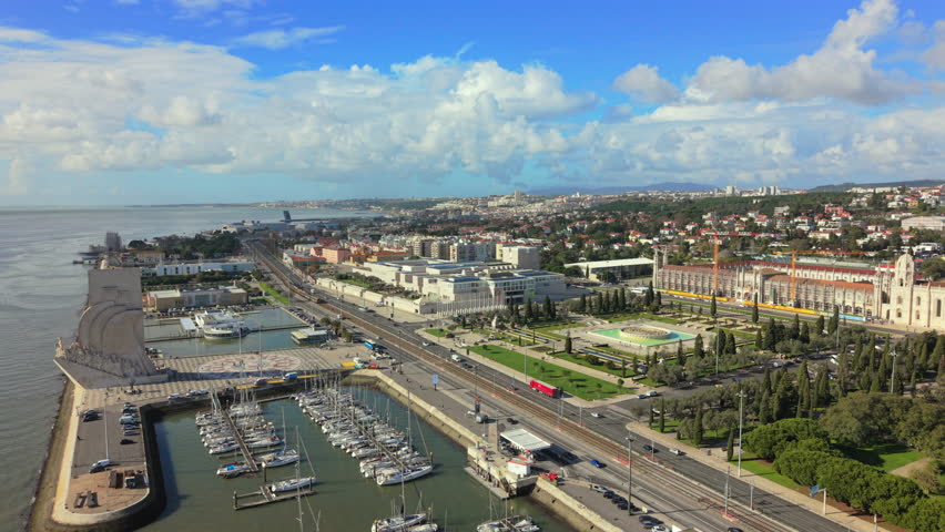 Aerial view of Lisbon Portugal showing Monument of the Discoveries, Ajuda Palace, marina with boats, riverfront promenade, coastal road and urban landscape under blue sky.