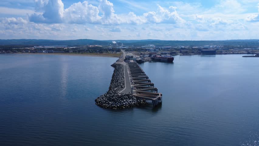 A long stone jetty juts out into a tranquil bay, connecting the shore to a bustling port. In the distance, warehouses and ships add an industrial touch to this peaceful seascape. Matane Port, Quebec, Canada, 2025.