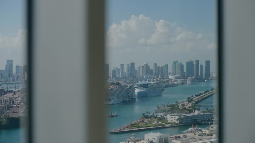 City view through vertical blinds shows waterfront with cruise ships and smaller boats, tall buildings rise behind. Green trees line promenade, sky partly cloudy