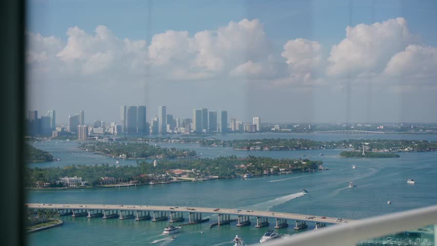 Coastal city view shows wide bridge crossing water, boats move below, skyline rises behind. Tall buildings stand near shore, partly cloudy sky with soft clouds adds depth