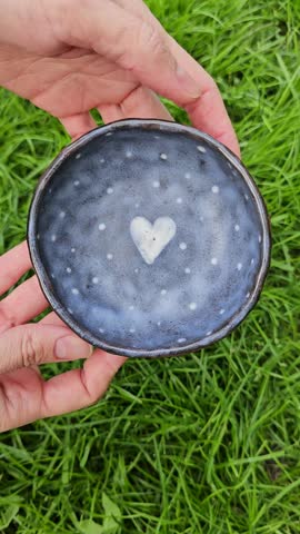 Small handcrafted ceramic bowl made of red clay with a simple heart motif inside. The video shows the bowl slowly rotating in hands, revealing its rounded shape, glazed interior, natural clay exterior, base, and subtle surface texture from all sides. Neutral background, suitable for handmade ceramics, tableware, gift, and lifestyle visuals.