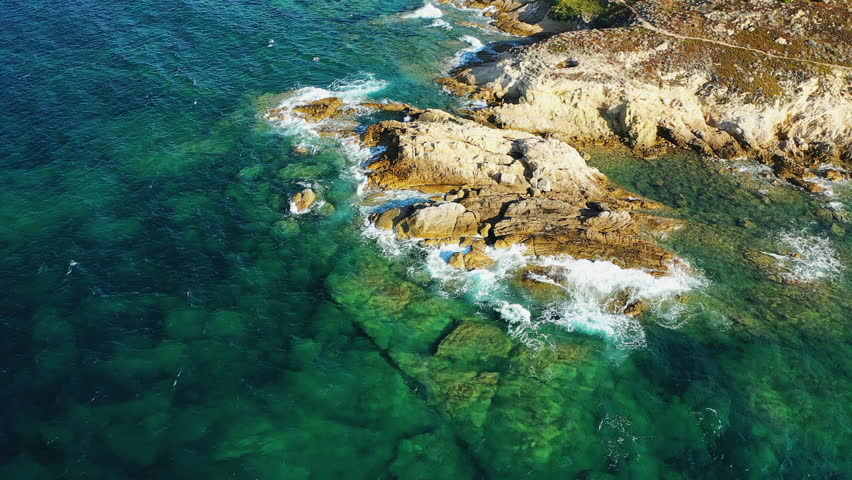 Aerial view of a rugged rocky coastline with clear turquoise water and waves crashing against the shore, Corsica, France.