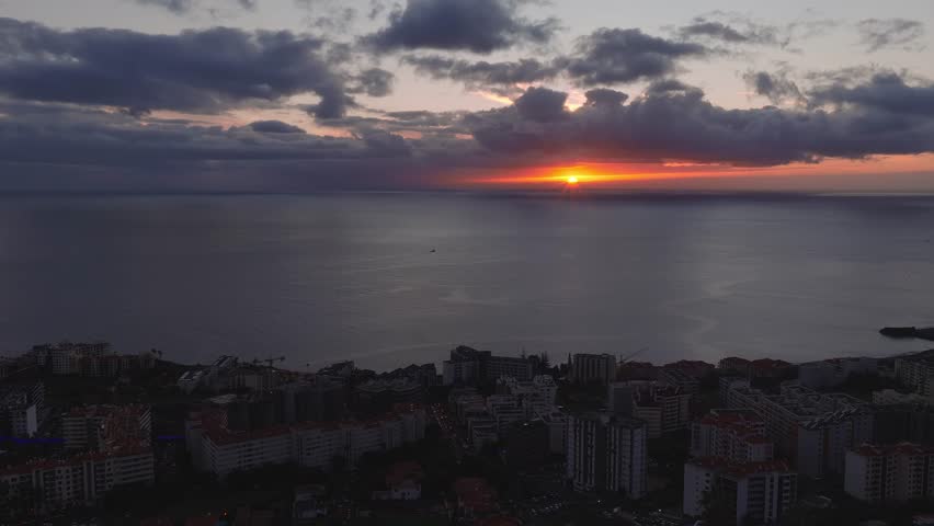 Aerial dusk view of Funchal, Madeira, Portugal, as a small boat moves past a dark headland, urban blocks line the coast, and low clouds reflect warm orange light.