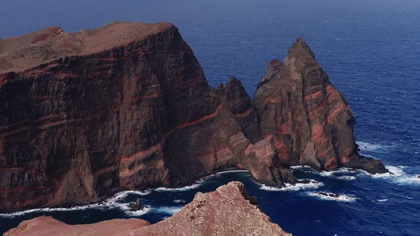 Aerial view shows red banded volcanic cliffs and a jagged sea stack at Ponta de Sao Lourenco, Madeira, Portugal, as waves churn below under daylight with high contrast.