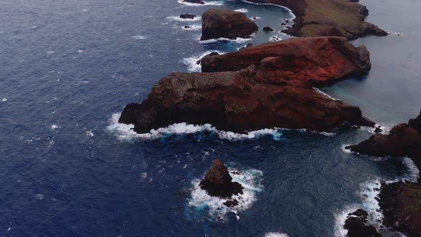 Aerial view shows Ponta de Sao Lourenco, Madeira, Portugal. Indigo Atlantic waves churn around sea stacks and coves as the camera sweeps slowly in overcast light.