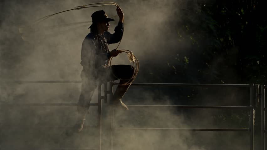A cowboy sitting on a corral fence skillfully spinning a lasso in dusty morning light