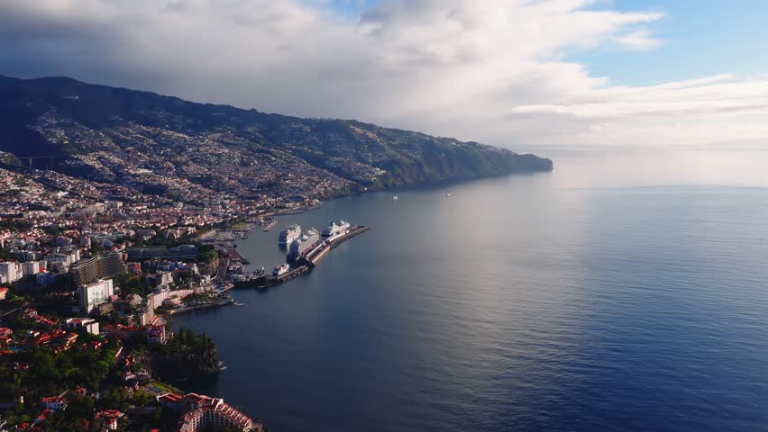 Aerial view of Funchal, Madeira, Portugal. Cruise liners dock, slow boats move, ripples form, clouds drift, sun glints on water, cliffs rise, piers and promenade stand.