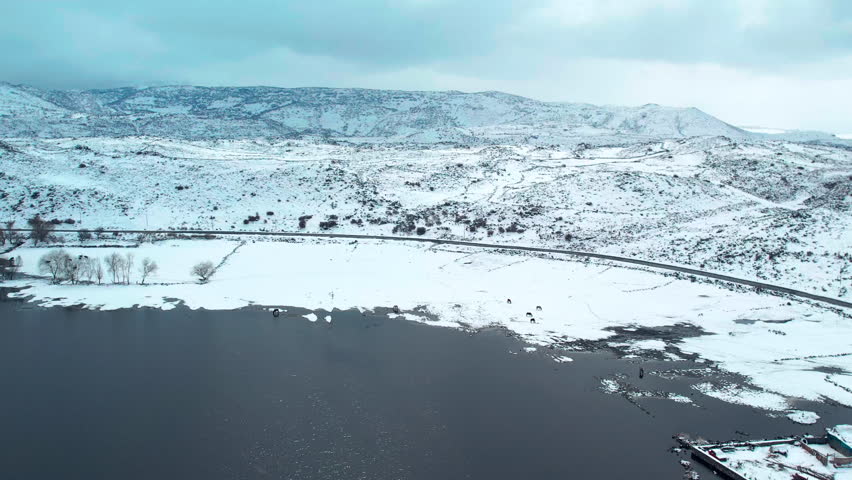 Wide aerial view of a calm lake surrounded by snow-covered land and distant hills. Minimal winter landscape showing cold climate, open water, natural patterns, and untouched nature.