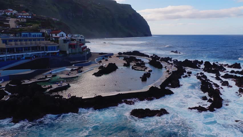 Drone view of Porto Moniz lava pools in Madeira. Waves hit breakwaters, swimmers use promenades and platforms. Turquoise pools contrast with dark basalt and ocean.