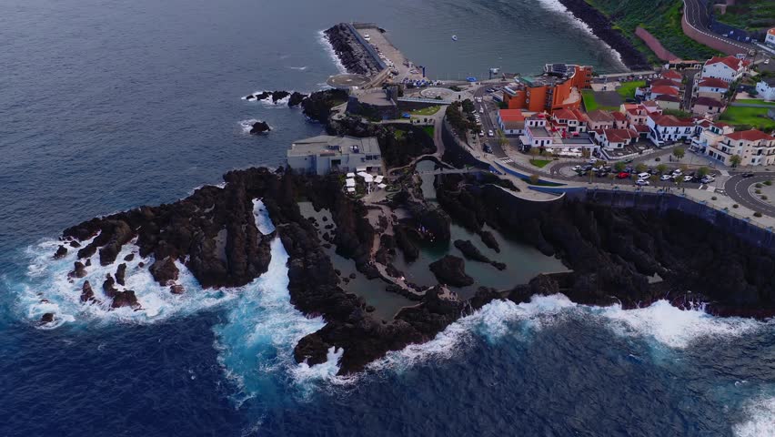 Aerial view shows Porto Moniz tidal pools, jagged basalt, harbor, and promenade as waves crash, boats move, and visitors walk beside red roof houses in Madeira.