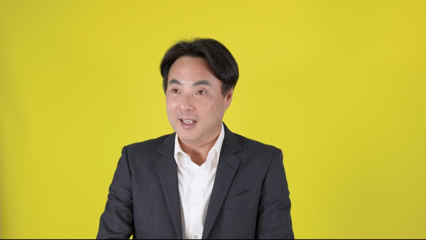 Businessman in suit smiles, gestures with hand, presenting product, brightly lit yellow studio background