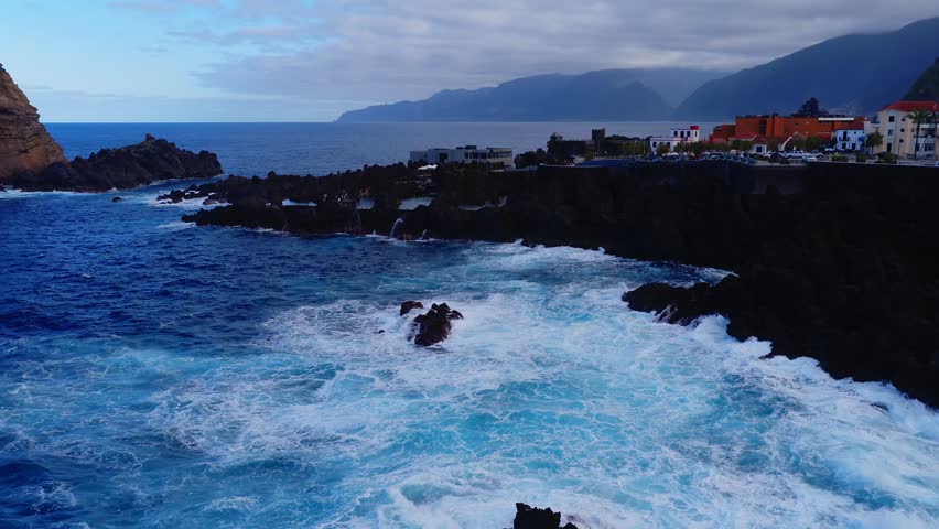 Aerial sweep shows Porto Moniz lava pools, promenade, and fortress like tower in Madeira. Late afternoon cool light, waves pound basalt, umbrellas reflect in pools.