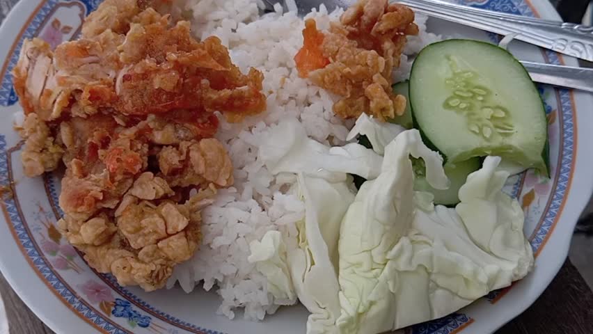 A plate of white rice with crispy fried chicken served with fresh vegetables in the form of cabbage and sliced cucumber.