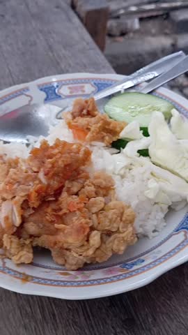 A plate of white rice with crispy fried chicken served with fresh vegetables in the form of cabbage and sliced cucumber.