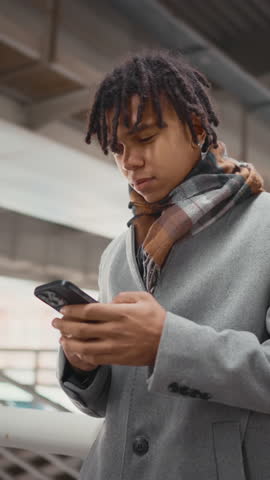 Young black man standing alone on city street and waiting someone, meeting in downtown. Vertical portrait of african american male person in coat looking around, date or gathering with friends