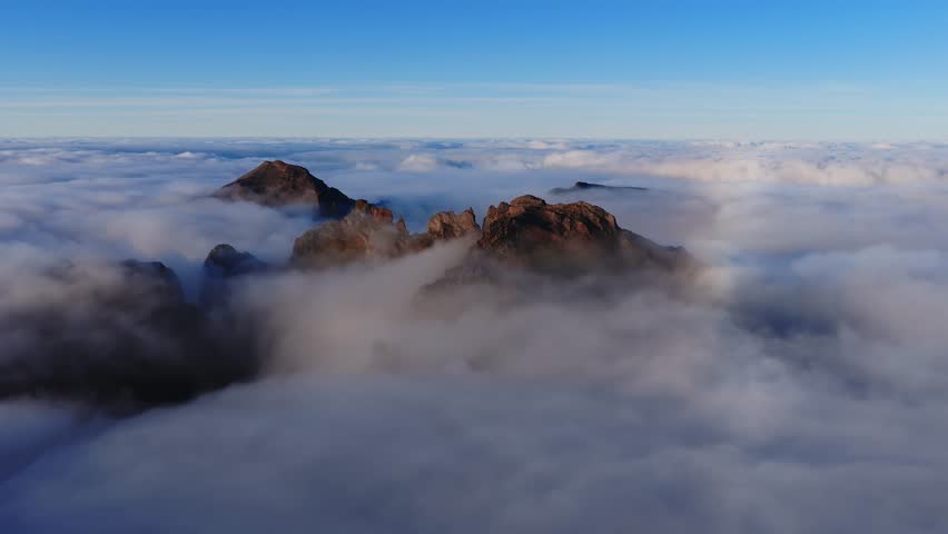 Rugged volcanic peaks in Madeira central highlands pierce clouds as early light warms rocky crags. Pico do Arieiro wide panoramic aerial view shows flowing mist and vertical relief.