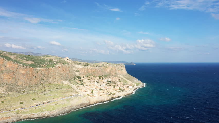 Scenic aerial view of the historic Monemvasia village nestled against dramatic cliffs and the deep blue Aegean Sea in Greece under a bright sky.