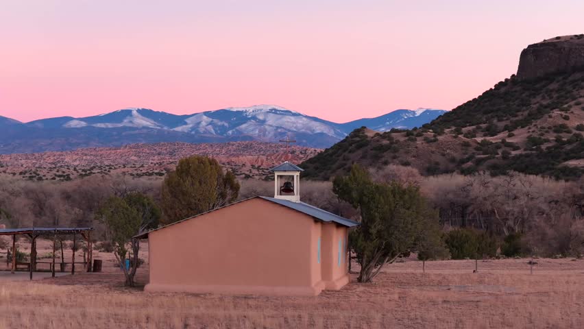 A cinematic drone orbit of a rural church with stunning light blue features at dusk. The shot circles to reveal snowy mountains and vast desert chaparral landscape.