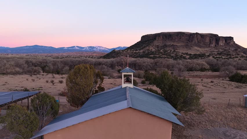 High-end drone establishing shot pulls back and orbits a rural church cross at dusk. Reveals a vast desert landscape with a large mesa and snow-capped mountains. Perfect for documentary openings.