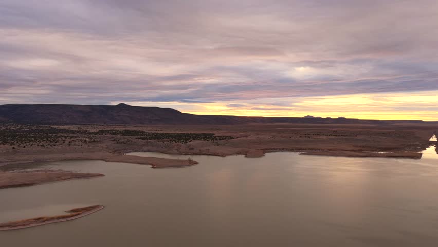 Professional drone establishing shot rises and rotates toward distant mountains over a serene desert lake at sunrise. Ideal for cinematic documentary openings featuring vast, rugged wilderness.