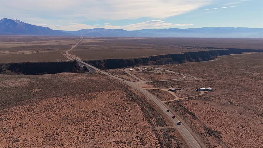 Cars drive along the straight desert road toward the Rio Grande Gorge Bridge. This wide view captures the bridge crossing the deep volcanic canyon and river in the New Mexico high desert.
