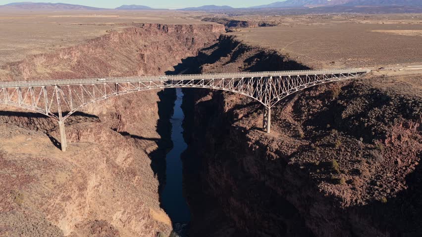 The Rio Grande Gorge Bridge crosses a deep volcanic canyon. This view shows cars on the steel bridge roadway above the Rio Grande River in the arid New Mexico high desert plateau landscape.