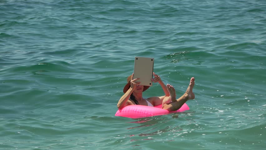 Woman Tablet Ocean. Woman floats on pink ring, raising tablet to capture sunny sea view on summer vacation.