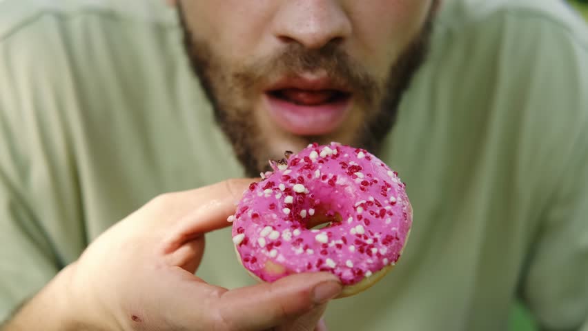 Young bearded man takes a big bite of a pink frosted donut with sprinkles outdoors, enjoying a fun sweet moment. Close up view portrait. 4k horizontal footage