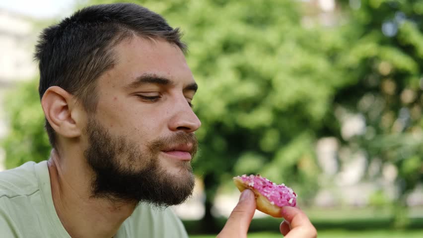 Young bearded man takes a big bite of a pink frosted donut with sprinkles outdoors, enjoying a fun sweet moment. Side view portrait. 4k horizontal footage