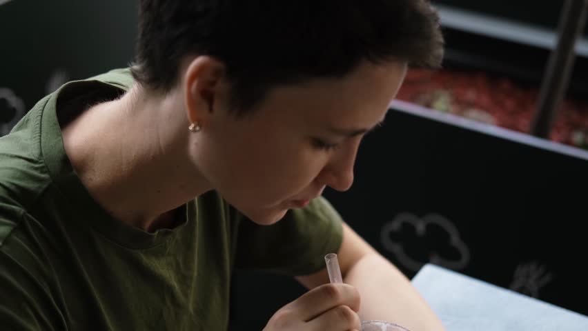 Young woman with short hair enjoys iced latte with whipped cream at an outdoor cafe on a warm day, relaxed and thoughtful moment. 4k horizontal footage