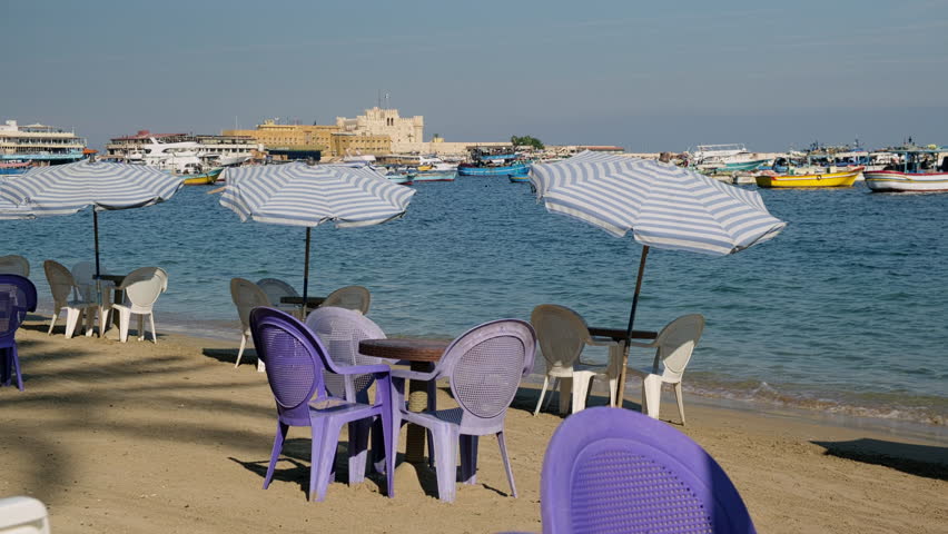 Sunny seaside scene with plastic chairs and tables arranged on sandy beach under striped umbrellas. Calm blue water, small boats and Qaitbay Fort in the background. Harbor of Alexandria, Egypt