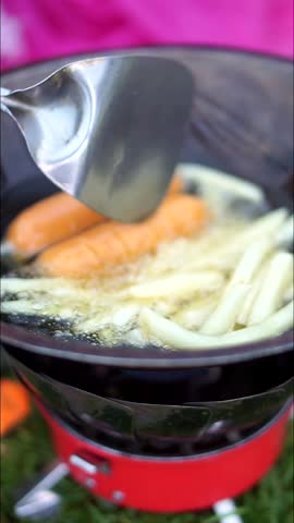 Frying potatoes and sausages in a pan filled with hot oil during outdoor cooking activity.