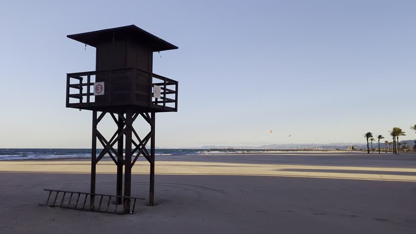 Lifeguard post on Cullera Beach, Valencia, Spain, with kitesurfers riding the waves and palm trees lining the Mediterranean coastline on a sunny and windy day.