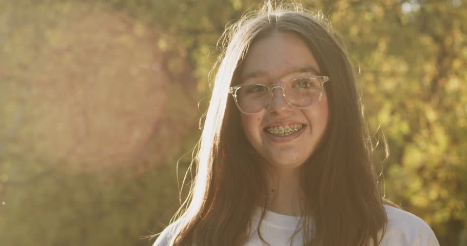 A portrait of a young girl wearing glasses, standing in the backlight of the sun and smiling at the camera, with metal braces visible on her teeth.