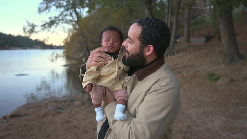 Father stands on a quiet lakeshore holding his infant, enjoying gentle daylight and peaceful surroundings while sharing a warm family moment in a soft relaxed natural atmosphere